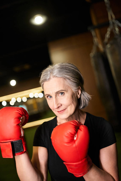 atraktivní starší veselá žena ve sportovním oblečení s šedivými vlasy pózování s boxerskými rukavicemi, zatímco v posilovně - Fotografie, Obrázek