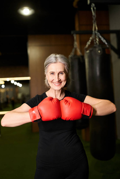 ελκυστική ώριμη χαρούμενη γυναίκα σε αθλητικά ρούχα με γκρίζα μαλλιά ποζάρουν με γάντια πυγμαχίας, ενώ στο γυμναστήριο - Φωτογραφία, εικόνα