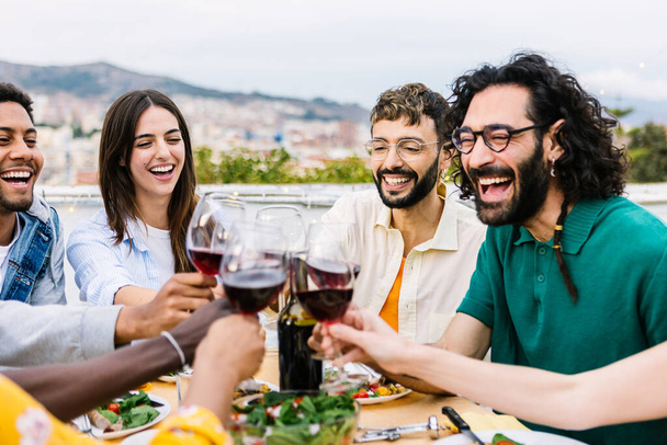 Ομάδα νέων που διασκεδάζουν πίνοντας κόκκινο κρασί στο πάρτι της ταράτσας. Πολυφυλετικές φίλες γιορτάζουν μαζί στο σπίτι βεράντα. - Φωτογραφία, εικόνα