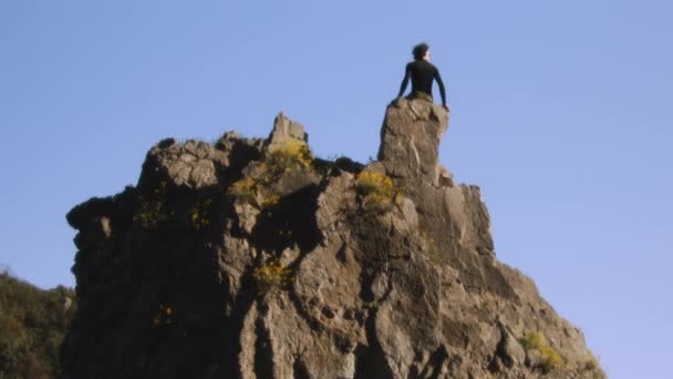 Un escalador se levanta victorioso en la cresta rugosa, brazos levantados en júbilo contra un cielo despejado. Esta toma cinematográfica, tomada con una cámara Blackmagic de alta calidad, congela el momento de triunfo - Imágenes, Vídeo