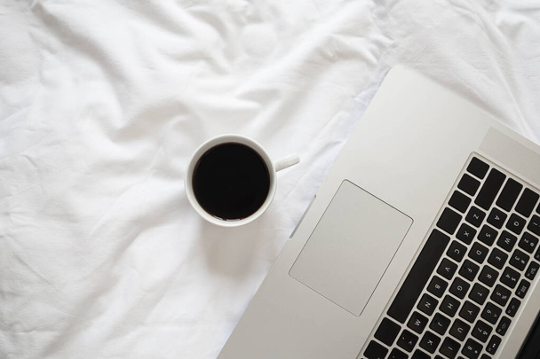 Laptop and coffee in bed on white linens, flat lay, top view minimalist workspace background, working at morning concept. - Photo, Image