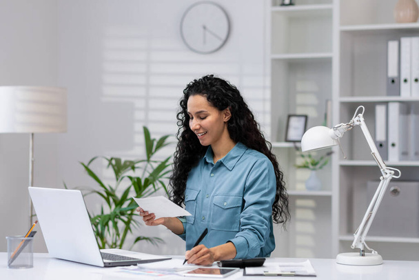 A cheerful Hispanic woman manages paperwork in her modern home office, exuding professionalism and positivity. - Photo, image