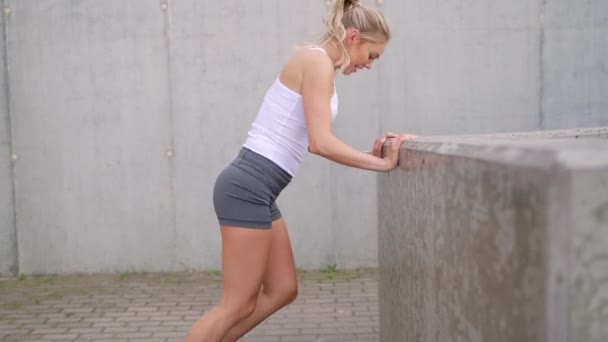 Jovem focada em sportswear alongamento por uma parede de concreto, preparando-se para uma rotina de fitness fora. - Filmagem, Vídeo