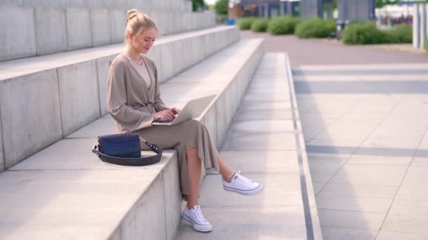 中立的な服装のファッショナブルな若い女性は,外で晴れた日を楽しんで,彼女のラップトップに意図的に取り組むステップに座っています. - 映像、動画