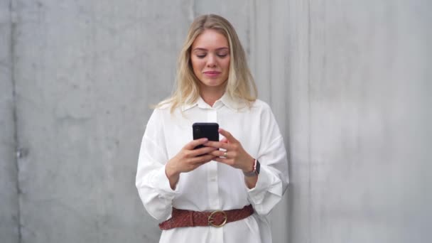Μια νεαρή ξανθιά γυναίκα με λευκή μπλούζα χρησιμοποιεί το smartphone της σε ένα γκρίζο αστικό σκηνικό, ενσωματώνοντας επαγγελματισμό και συνδεσιμότητα. - Πλάνα, βίντεο