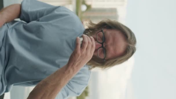 Vertical zoom-in portrait of adult Caucasian man with long hair, beard and glasses looking at camera while drinking hot tea from thermos cup sitting outside by water in city park - Footage, Video