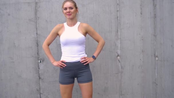 Past jonge vrouw in sportkleding staat vol vertrouwen tegen een betonnen muur, het nemen van een pauze na een oefening sessie. - Video