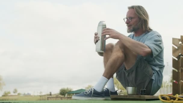 Hombre adulto caucásico sentado en la estera de césped verde artificial al aire libre y beber té caliente de termos - Metraje, vídeo