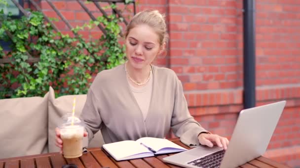 Soustředěný mladý businesswoman pomocí svého notebooku v útulné venkovní kavárně, popíjení nápoj přes brčko. - Záběry, video