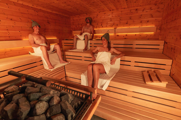 Tři lidé v sauně ve finských plstěných čepicích, dva muži sedící a žena ležící - Fotografie, Obrázek