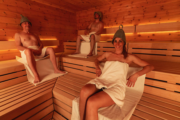 Saunada üç kişi keçeli şapka takıyor, iki erkek oturuyor ve bir kadın yaslanıyor. - Fotoğraf, Görsel