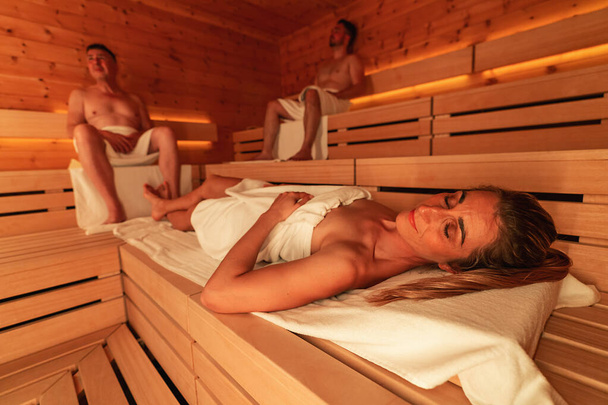 Mujer reclinada y hombres sentados en una sauna finlandesa de madera, luz ambiental cálida - Foto, imagen
