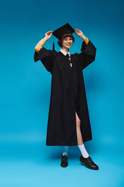 happy college girl wearing black graduation gown and cap standing on blue background in studio - Photo, Image