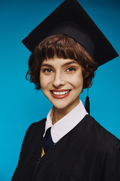 retrato de chica universitaria sonriente con vestido de graduación negro y gorra académica sobre fondo azul - Foto, imagen