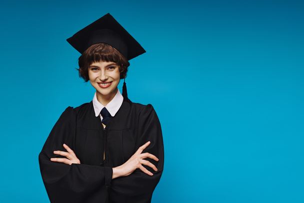 Porträt eines lächelnden College-Mädchens in schwarzem Abschlusskleid und akademischer Mütze, das mit verschränkten Armen posiert - Foto, Bild