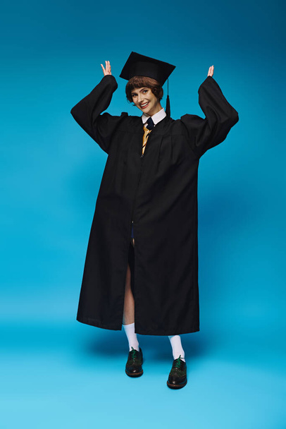 graduation concept, cheerful college girl in academic cap and gown standing on blue background - Photo, Image