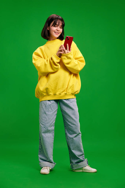 Retrato completo de mulher jovem em roupa de moda de tamanho grande usando smartphone e sorrindo contra o fundo do estúdio verde vibrante. Conceito de emoções humanas, moda, beleza, estilo. - Foto, Imagem