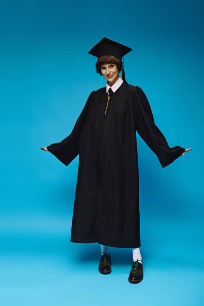 érettségi koncepció, optimista főiskolai lány akadémiai sapkában és köntösben álló kék háttér - Fotó, kép