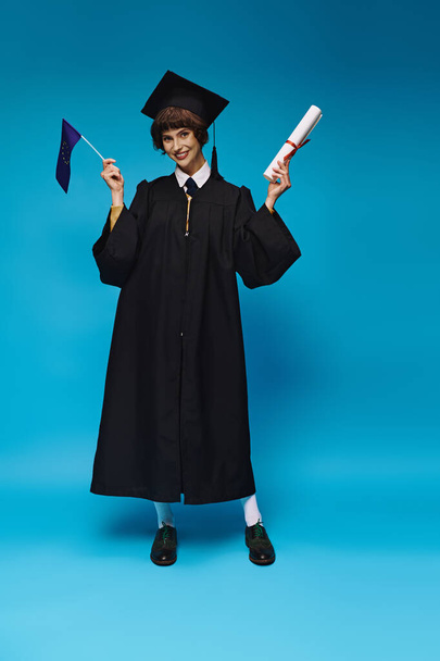 radosna absolwentka koledżu w sukni i czapce akademickiej z dyplomem i flagą UE na niebieskim tle - Zdjęcie, obraz