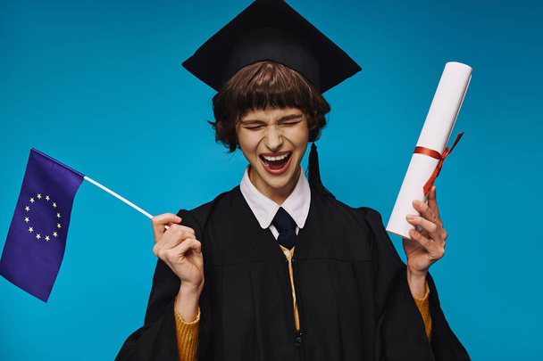 vzrušená vysokoškolačka v šatech a akademické čepici s diplomem a vlajkou EU na modrém pozadí - Fotografie, Obrázek
