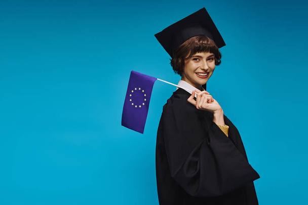 graduated college girl in gown and academic cap with diploma and EU flag smiling on blue backdrop - Photo, Image