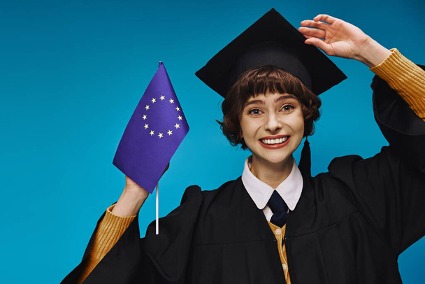 absolwentka w sukni i czapce z flagą UE i uśmiechnięta na niebieskim tle, edukacja europejska - Zdjęcie, obraz