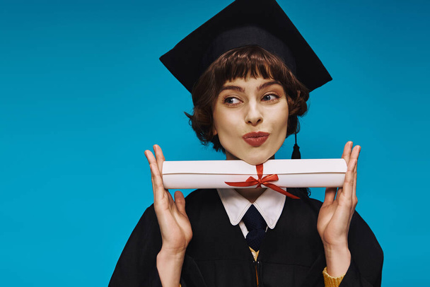 menina da faculdade grad feliz no vestido e tampão acadêmico lábios bebendo e segurando seu diploma com orgulho - Foto, Imagem