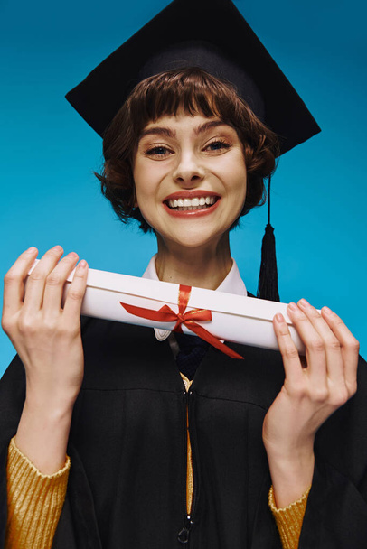 весела дівчина в коледжі в сукні і академічна шапка, дивлячись на камеру з дипломом в руках на синьому - Фото, зображення