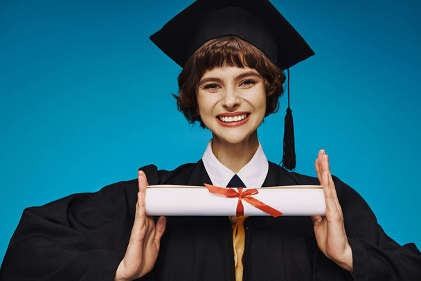 Mavi elbisesi ve akademik şapkası olan liseli kız diplomasını gururla tutuyor. - Fotoğraf, Görsel