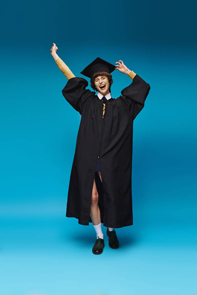 aufgeregtes Hochschulmädchen in Kleid und Mütze, das die Fäuste auf blauem Hintergrund hebt, Erfolg - Foto, Bild