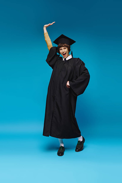excitado graduado chica universitaria en vestido y gorra levantamiento y y gritando de alegría en el fondo azul - Foto, imagen