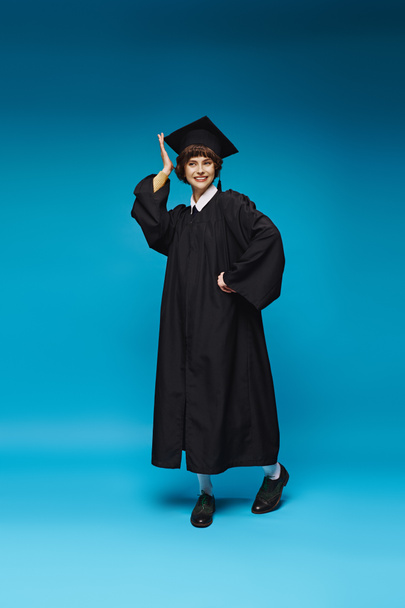 aufgeregtes Hochschulmädchen in Kleid und Mütze, lächelnd auf blauem Hintergrund, Vollendung - Foto, Bild