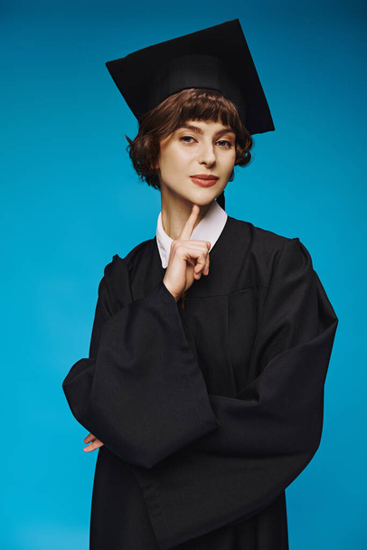 young graduated college girl in gown and cap standing on blue background, accomplishment - Photo, Image