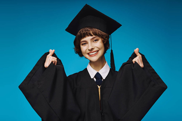 fröhliches Hochschulmädchen in akademischer Mütze, das mit den Fingern auf blauem Hintergrund Herzzeichen macht - Foto, Bild