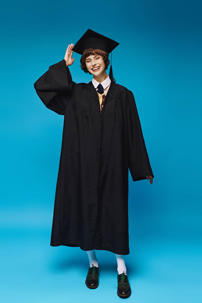 excité jeune femme en robe de graduation noire et chapeau académique souriant sur fond bleu, cérémonie - Photo, image