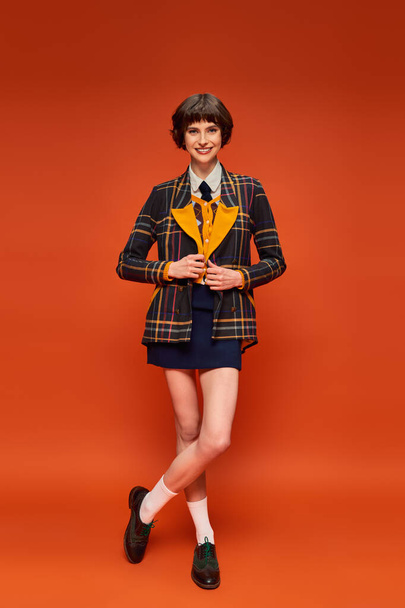 полная длина стильный студент в клетчатом блейзере и юбке на оранжевом фоне, униформа колледжа - Фото, изображение
