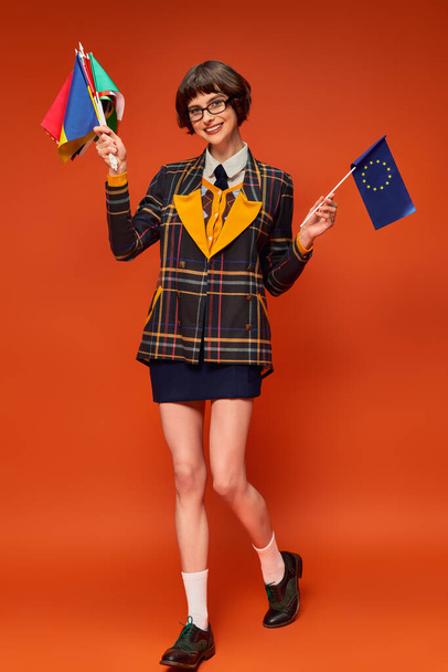 joven universitaria feliz en uniforme y gafas que sostienen la UE y diferentes banderas sobre fondo naranja - Foto, Imagen