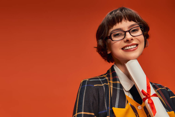 positive college girl in uniform and glasses holding her graduation diploma on orange backdrop - Photo, Image