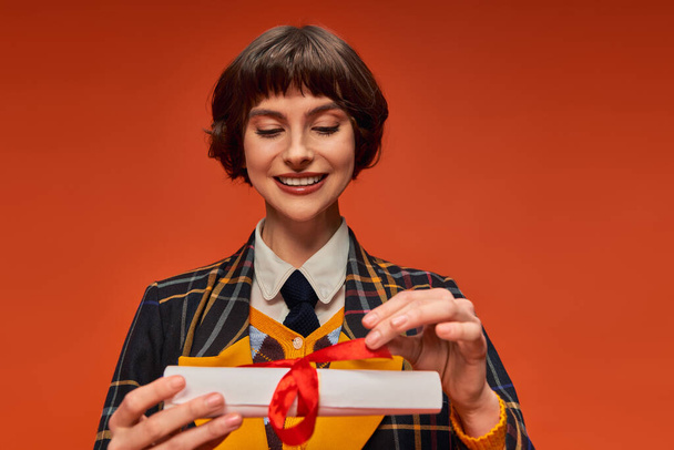portrait of happy college girl in checkered uniform looking at graduation diploma on orange backdrop - Photo, Image