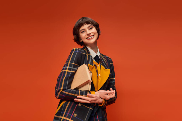 ritratto di studente felice in uniforme universitaria in piedi con libri su sfondo arancione, conoscenza - Foto, immagini