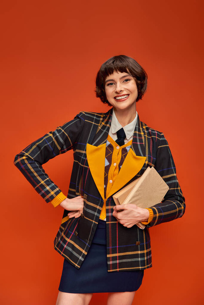 портрет улыбающегося студента в униформе колледжа, стоящего с книгами на оранжевом фоне, знания - Фото, изображение