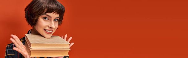 banner of smiling college girl in uniform holding books near chin on orange background, knowledge - Photo, Image