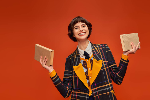 Porträt eines fröhlichen College-Mädchens in Uniform mit Büchern in der Hand auf orangefarbenem Hintergrund, Wissen - Foto, Bild