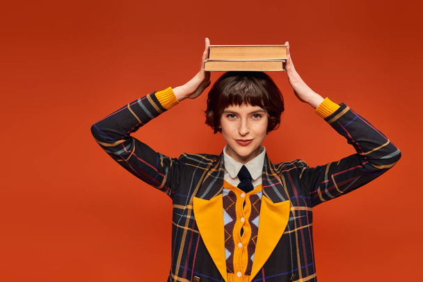 happy college girl in uniform holding stack of books on hand on orange background, knowledge - Photo, Image