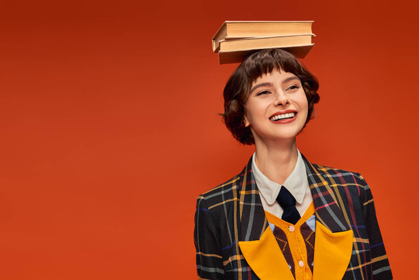 Üniformalı iyimser üniversiteli kız elinde turuncu arka planda bir yığın kitap tutuyor. - Fotoğraf, Görsel