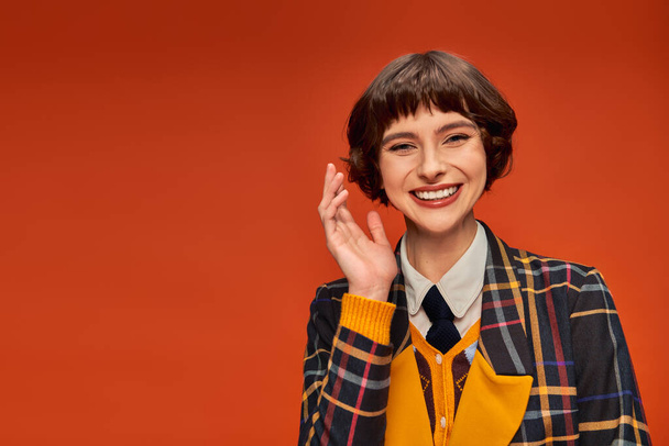optimistické vysokoškolačka v kostkované uniformě mávání rukou na oranžovém pozadí, šťastný studentský život - Fotografie, Obrázek