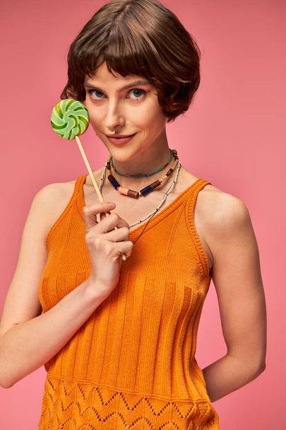 brunette young woman in orange knitted tank top holding colorful sweet lollipop on pink backdrop - Photo, Image
