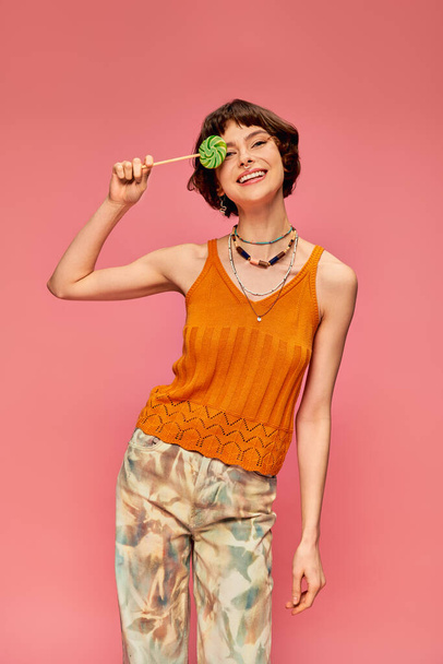happy young woman in orange knitted tank top covering eye with sweet lollipop on pink backdrop - Photo, Image