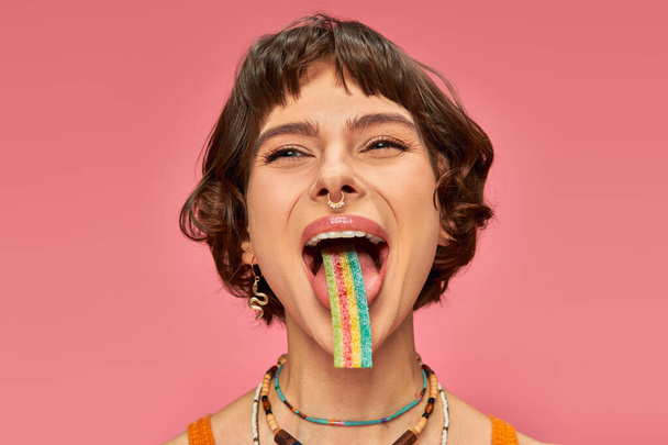 cheerful young woman in her 20s tasting sweet and sour candy strip on her tongue, pink background - Photo, Image