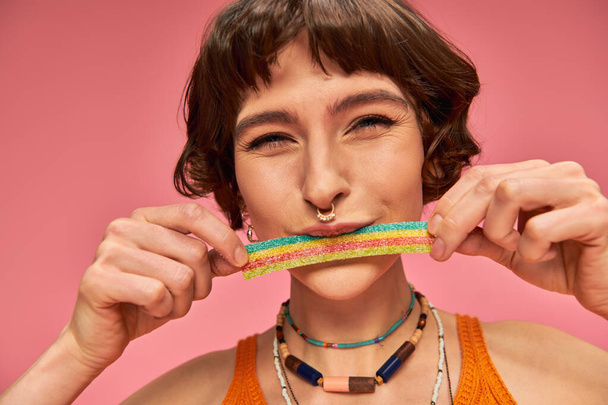 portrait of cheerful young woman in her 20s tasting sweet and sour candy strip on pink background - Photo, Image
